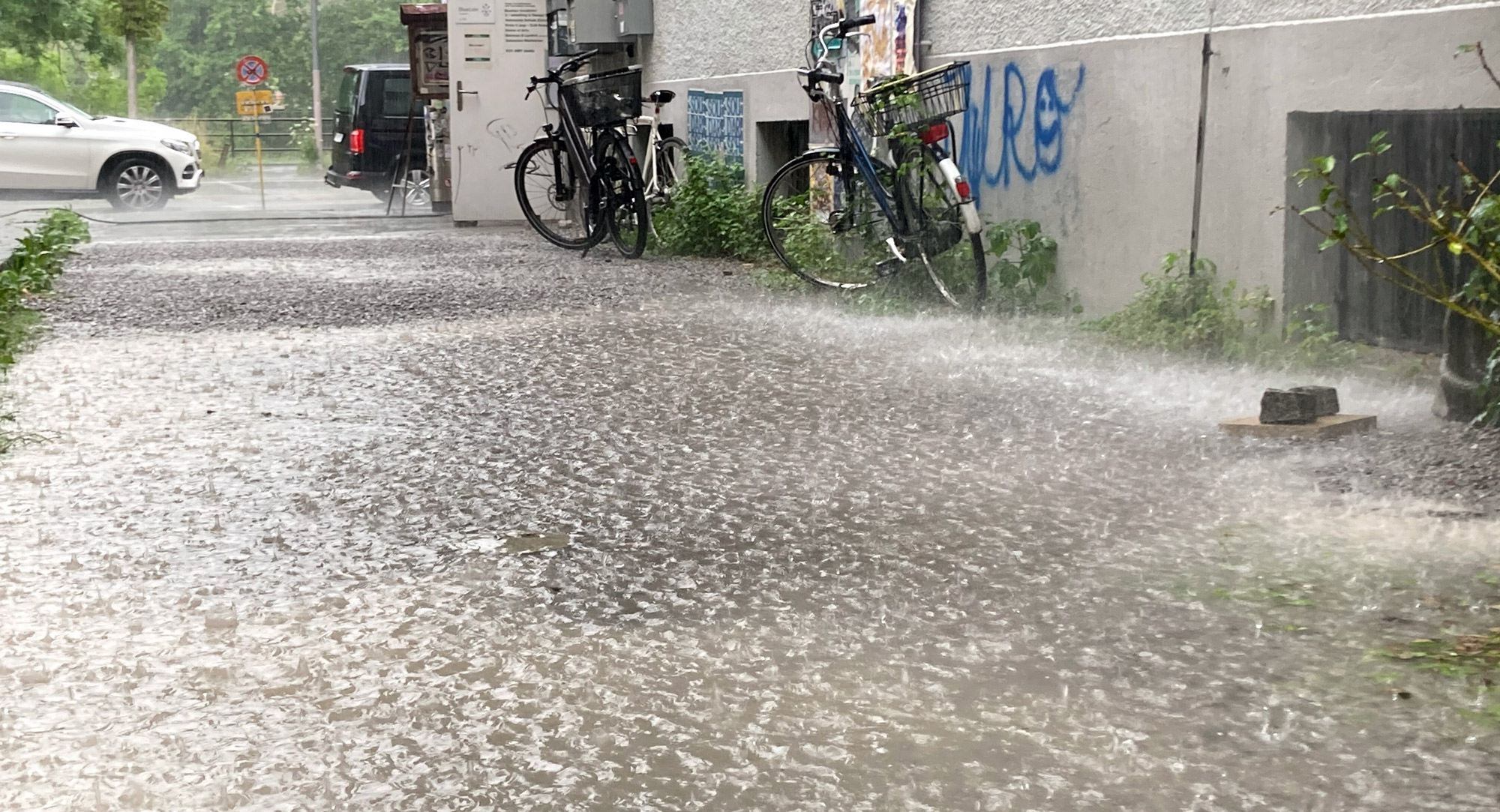 Eine überschwemmte Straße bei starkem Regen mit Fahrrädern, die an ein Gebäude gelehnt sind.