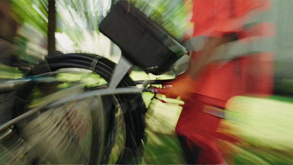 Unscharfes Bild einer Person in orangefarbener Kleidung, die im Freien in einer grünen, belaubten Umgebung ein Fahrrad mit einem Korb und auf der Rückseite aufgerollten schwarzen Kabeln schiebt.