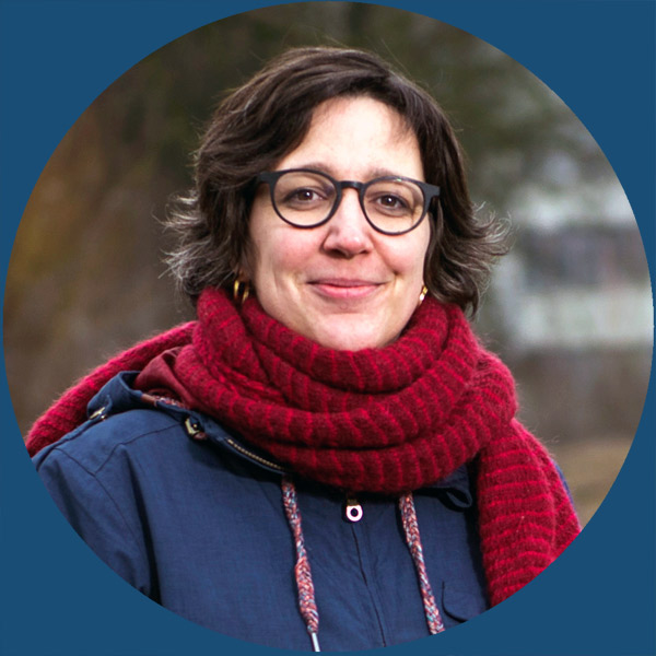 Eine Frau mit Brille, einer blauen Jacke und einem roten Schal lächelt im Freien.