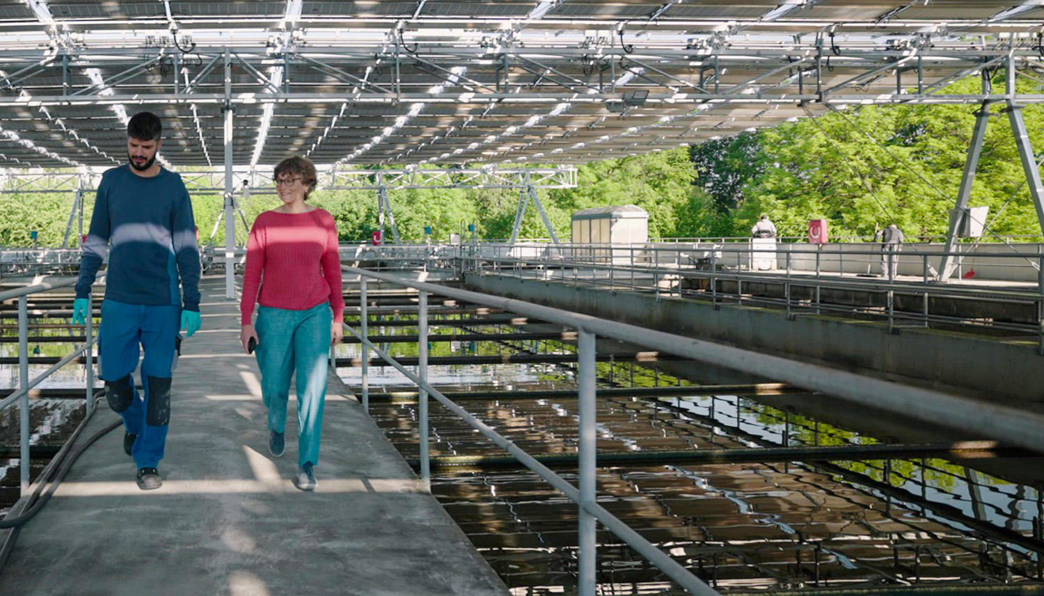Zwei Personen gehen auf einem Betonsteg im Inneren einer Wasseraufbereitungsanlage, umgeben von Metallgeländern und großen Tanks unter einer Dachkonstruktion.