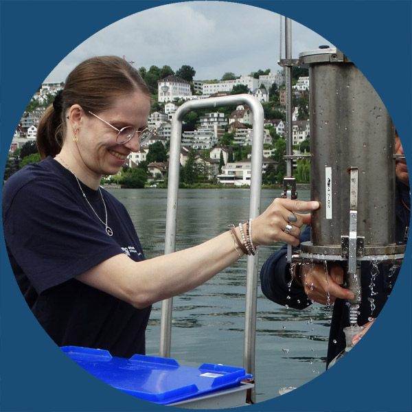Eine Person lächelt, während sie mit einer wissenschaftlichen Ausrüstung auf einem Boot hantiert. Im Hintergrund sind Häuser zu sehen, von denen Wasser auf das Gerät tropft.