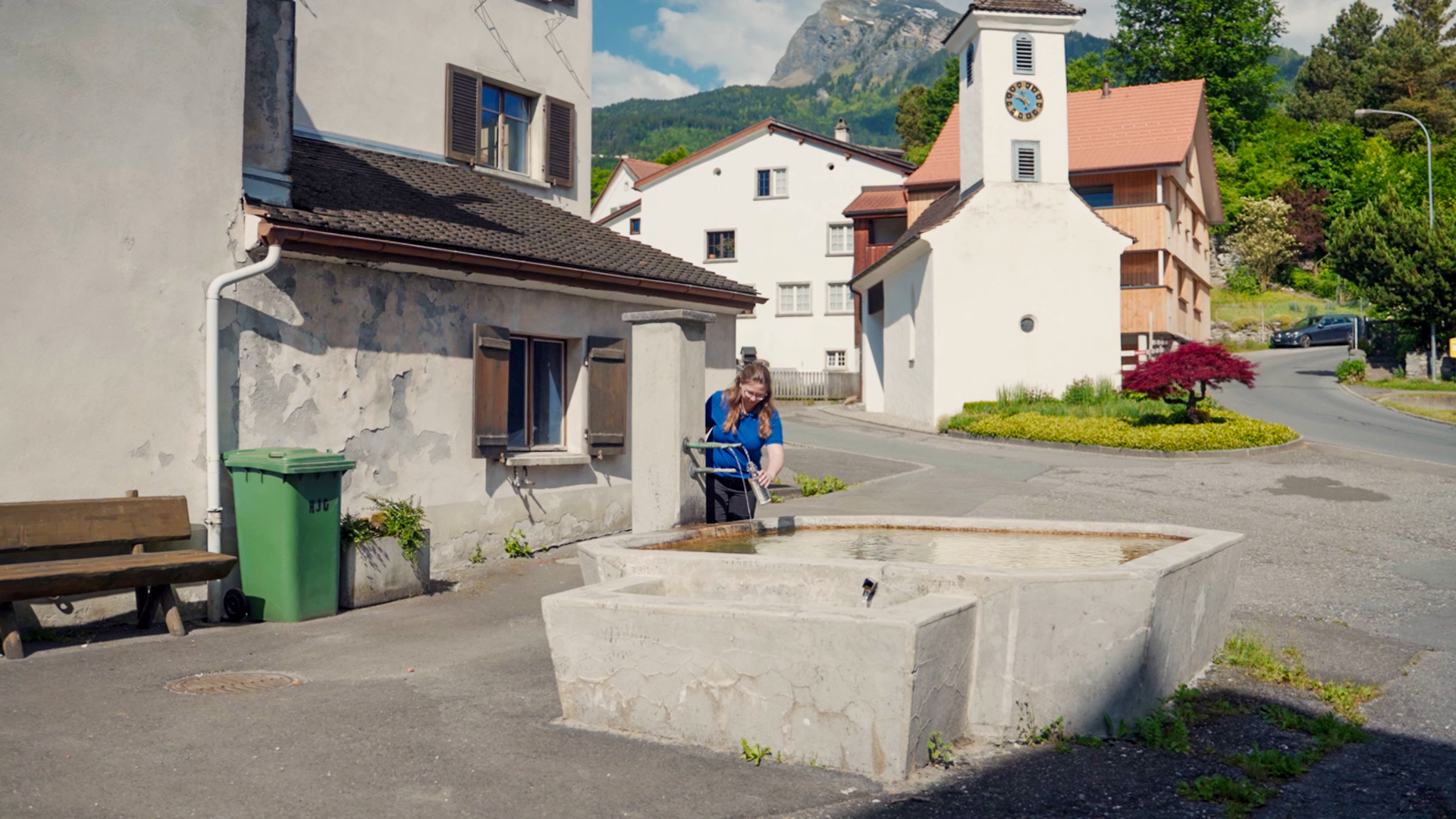 Eine Person füllt eine Flasche an einem steinernen Wasserbrunnen auf einem kleinen Dorfplatz mit traditionellen Häusern und einem Kirchturm im Hintergrund.